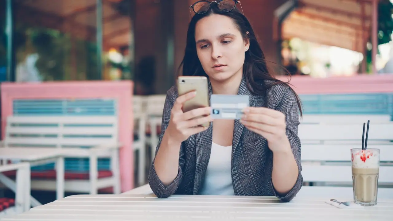 Donna riflessiva con in mano una carta di credito mentre guarda lo smartphone per fare acquisti online
